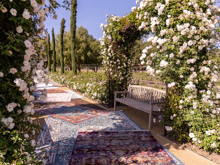 Entrance to Mizel Estate featuring a floral-encrusted bridge, a tree-lined path with rose bushes, and colorful Belgian rugs creating a romantic and elegant atmosphere.